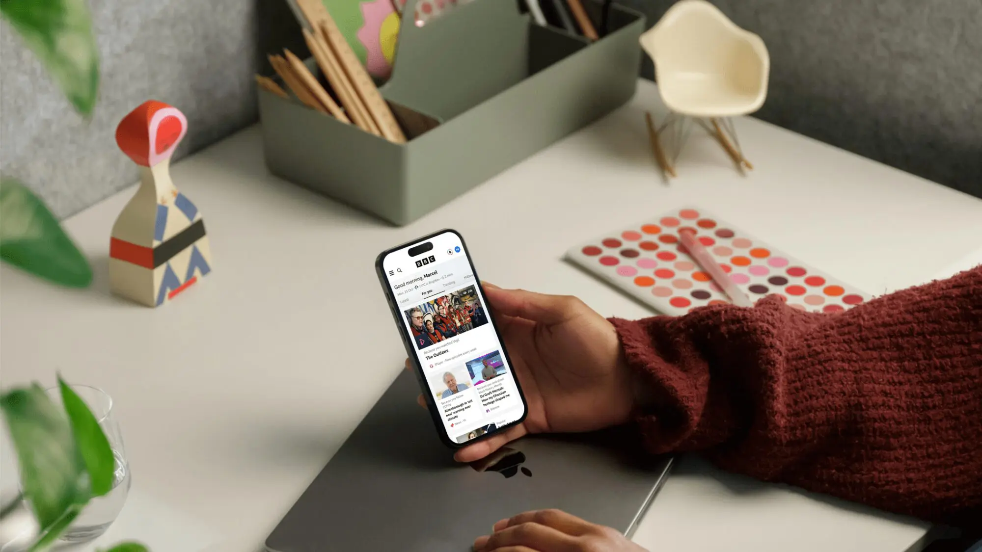 A hand rests on a desk holding a mobile phone with the new-look BBC homepage featured on it.