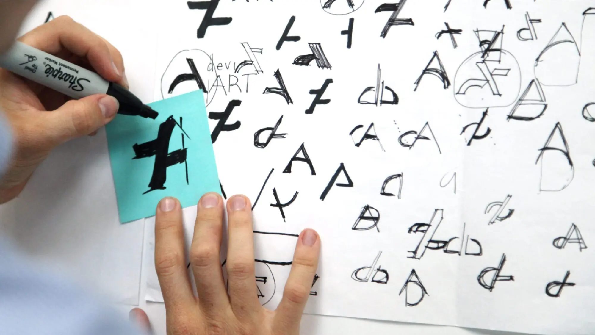 A close-up of hands sketching the A of the DeviantArt symbol, with multiple versions in the background.