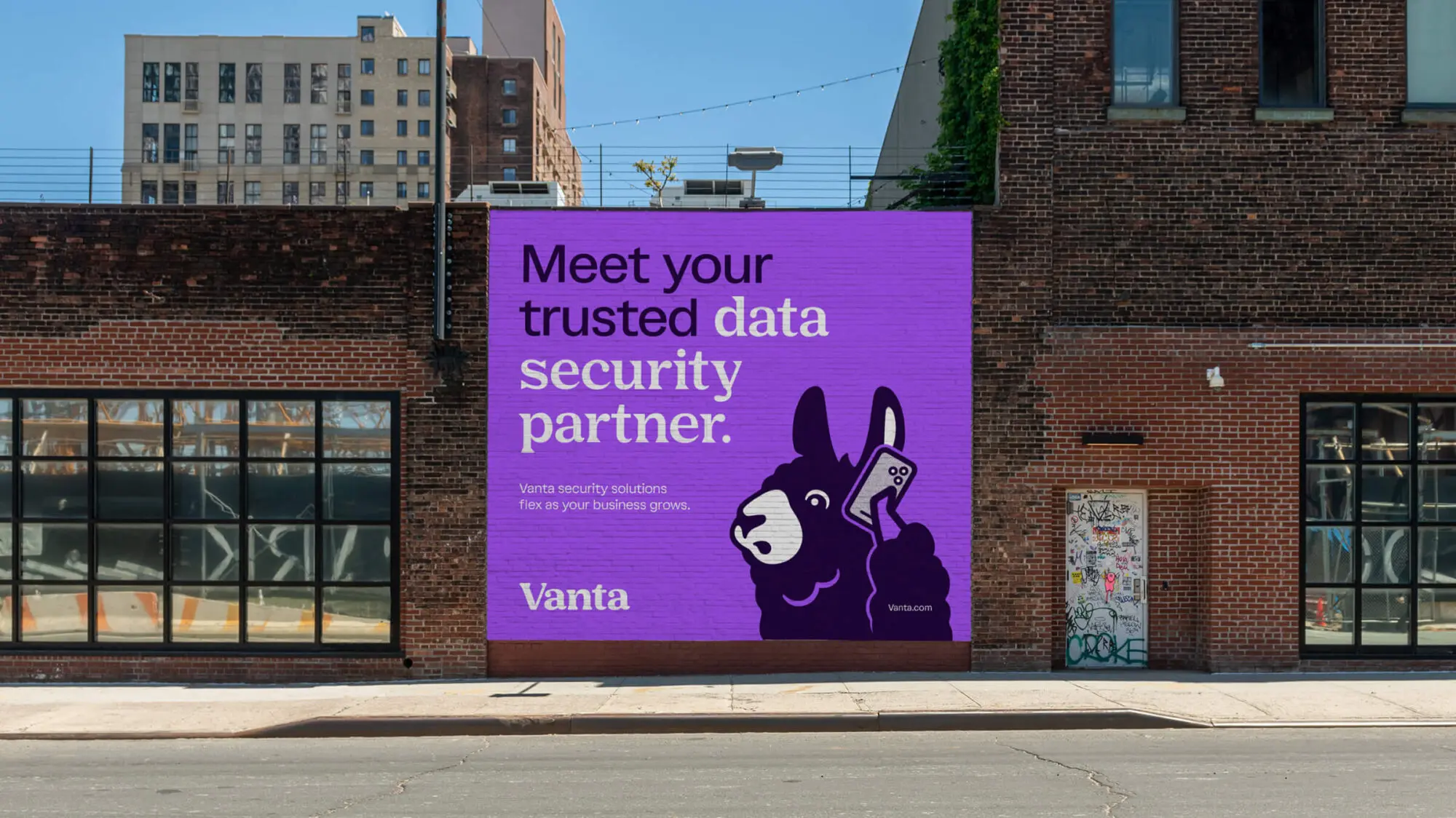 A street with a graphic Vanta poster pasted on the wall. The poster features a llama, the brand mascot, on the phone and the title “Meet you trusted data security partner”.